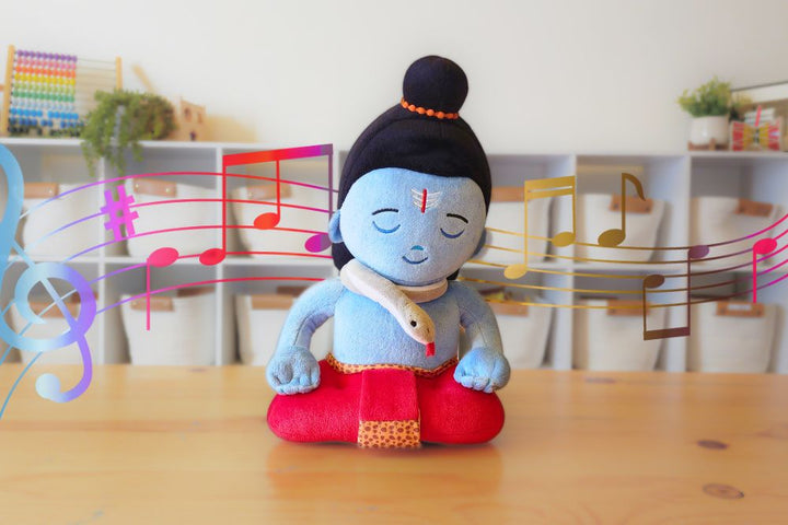 A plush toy depicting Lord Shiva in a meditation pose with closed eyes, wearing a blue outfit and a red cloth around the waist. The toy appears to be about 11 inches tall and is shown with musical notes in the background, indicating it might sing mantras.