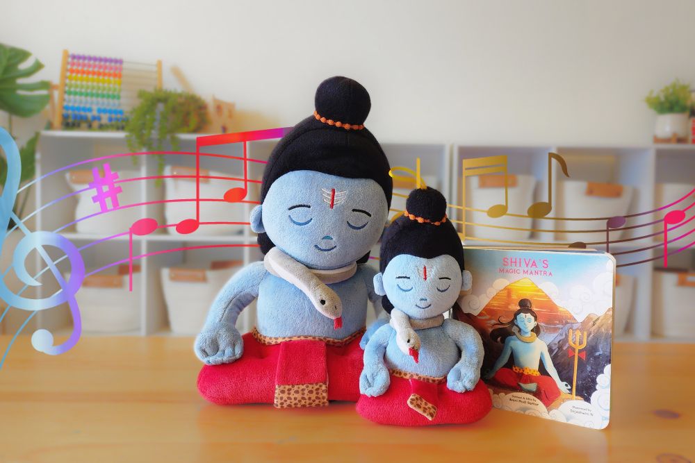 Two plush toys resembling the Hindu deity Shiv Shankar, one larger and one smaller, with a illustrated book in the background featuring the same deities. The toys appear to be multicolored with a predominantly blue and red outfit, and are posed with a music-related backdrop.